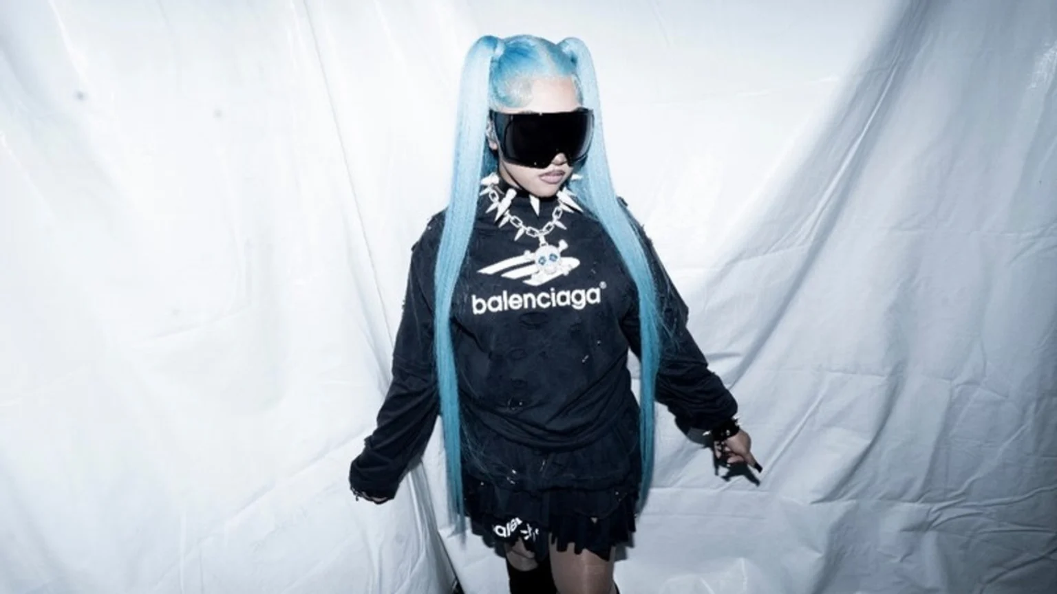 A person with light blue pigtails and large dark sunglasses wears a distressed black Balenciaga top, a spiked necklace with a skull pendant, and a black skirt against a white draped background.