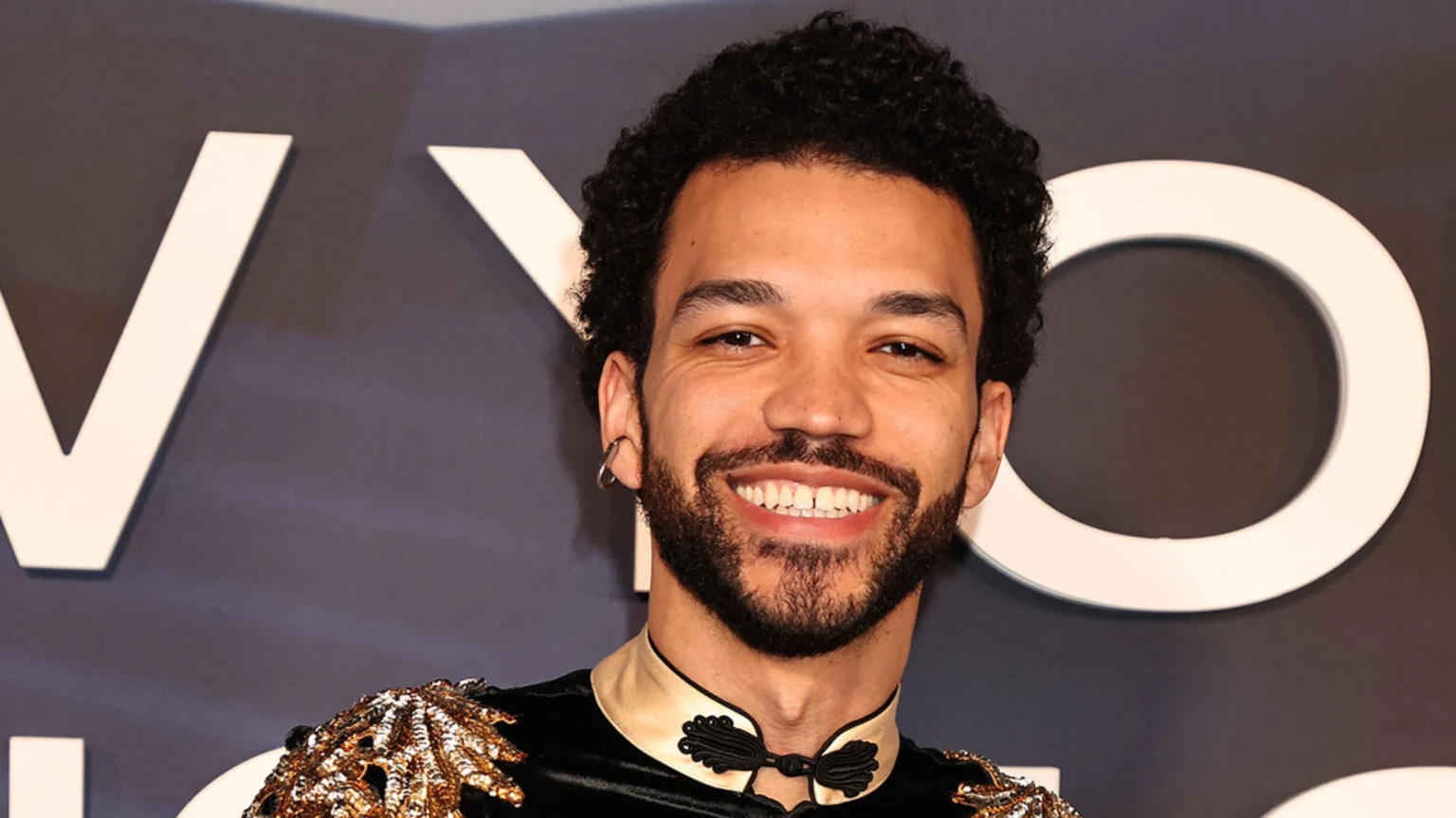Justice Smith, a man with curly hair and a beard, smiles broadly at the camera while wearing a black and gold ornate top, with large white letters visible in the background.