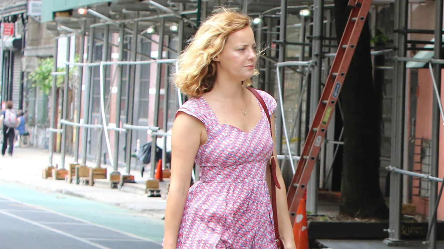 A woman with blonde curly hair, wearing a pink patterned dress and a red shoulder bag, walks past construction scaffolding and a red ladder on a city street.