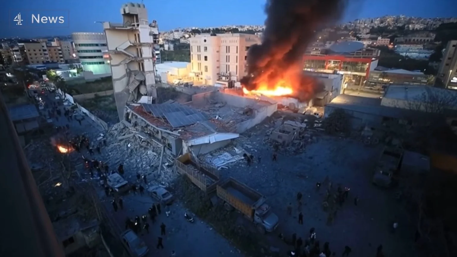 Aerial view of a city at dusk with a collapsed building and a large fire emitting thick black smoke, surrounded by debris, vehicles, and onlookers.