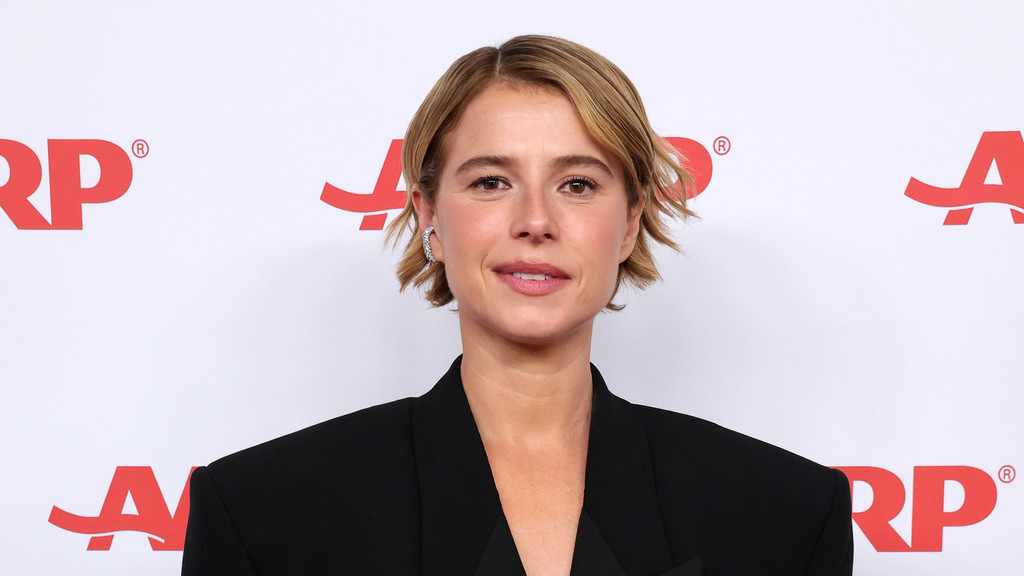 Jessie Buckley, a woman with short brown hair, looks directly at the camera, wearing a black blazer against a white background with red AARP logos.