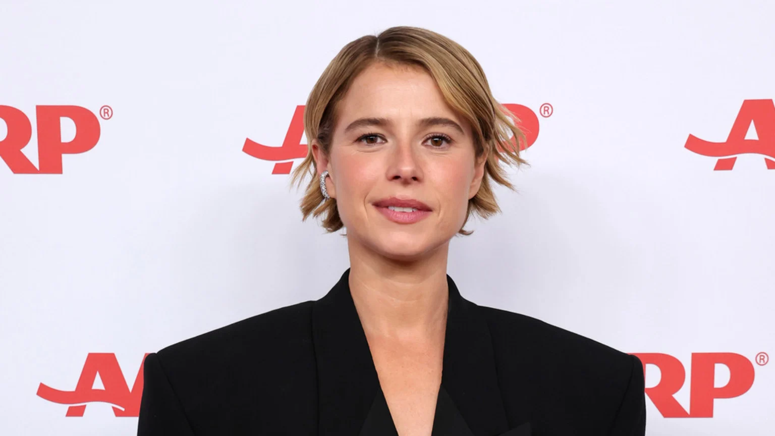 Jessie Buckley, a woman with short brown hair, looks directly at the camera, wearing a black blazer against a white background with red AARP logos.