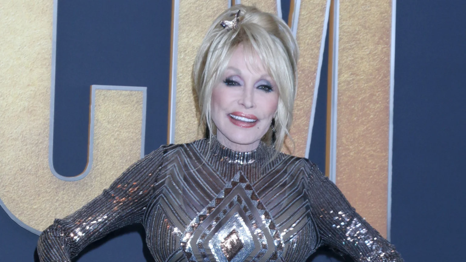 Dolly Parton smiles, wearing a sparkly, geometric patterned top with a high neck, against a dark blue and gold textured background with large, stylized letters.