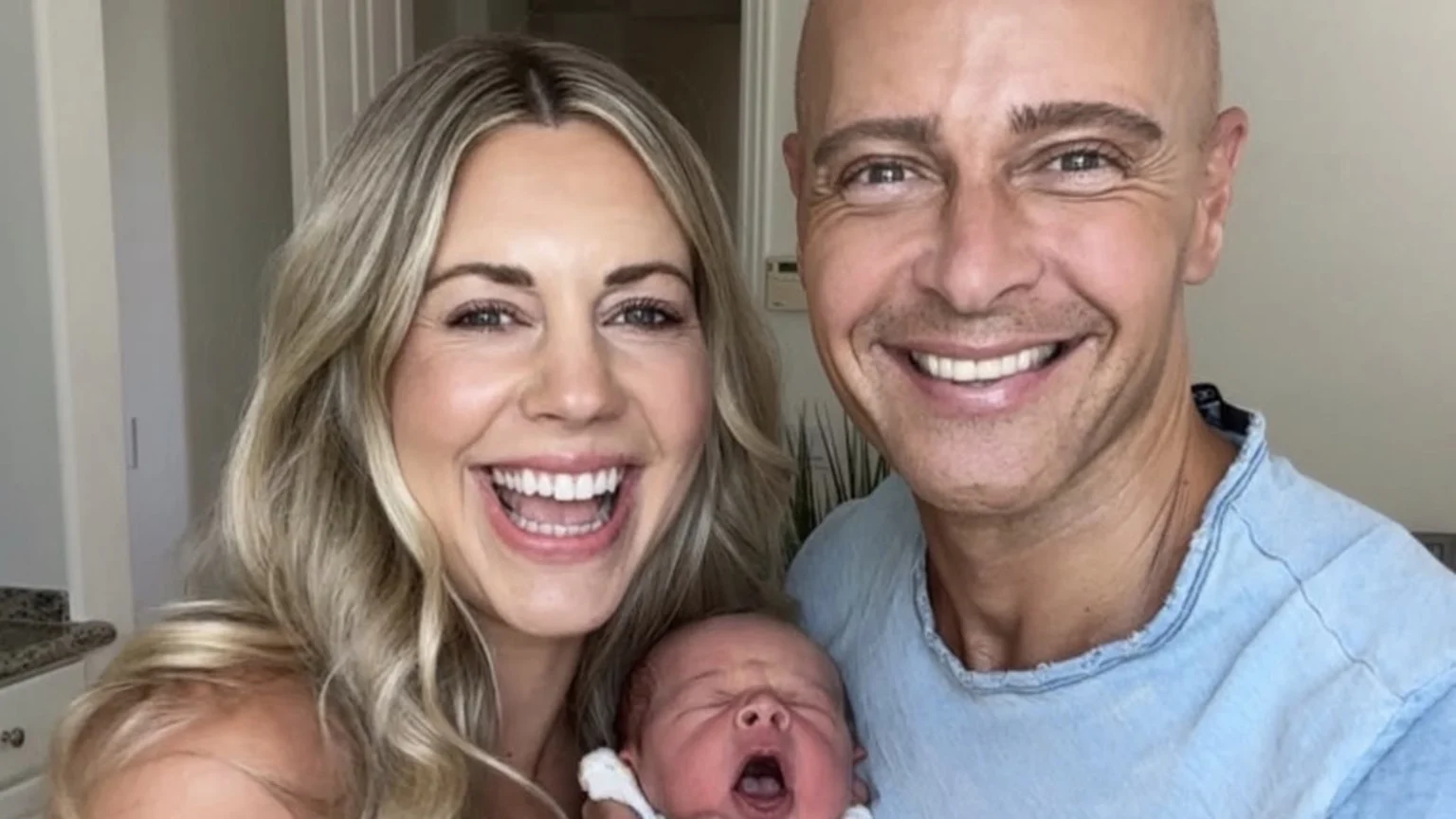 A smiling blonde woman and a bald man pose with a yawning newborn baby.