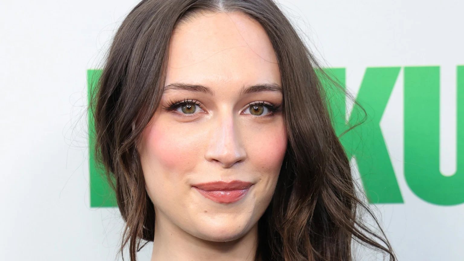 Close-up portrait of Elsie Hewitt with long brown hair, green-brown eyes, and pink makeup, smiling slightly against a white background with a green logo.