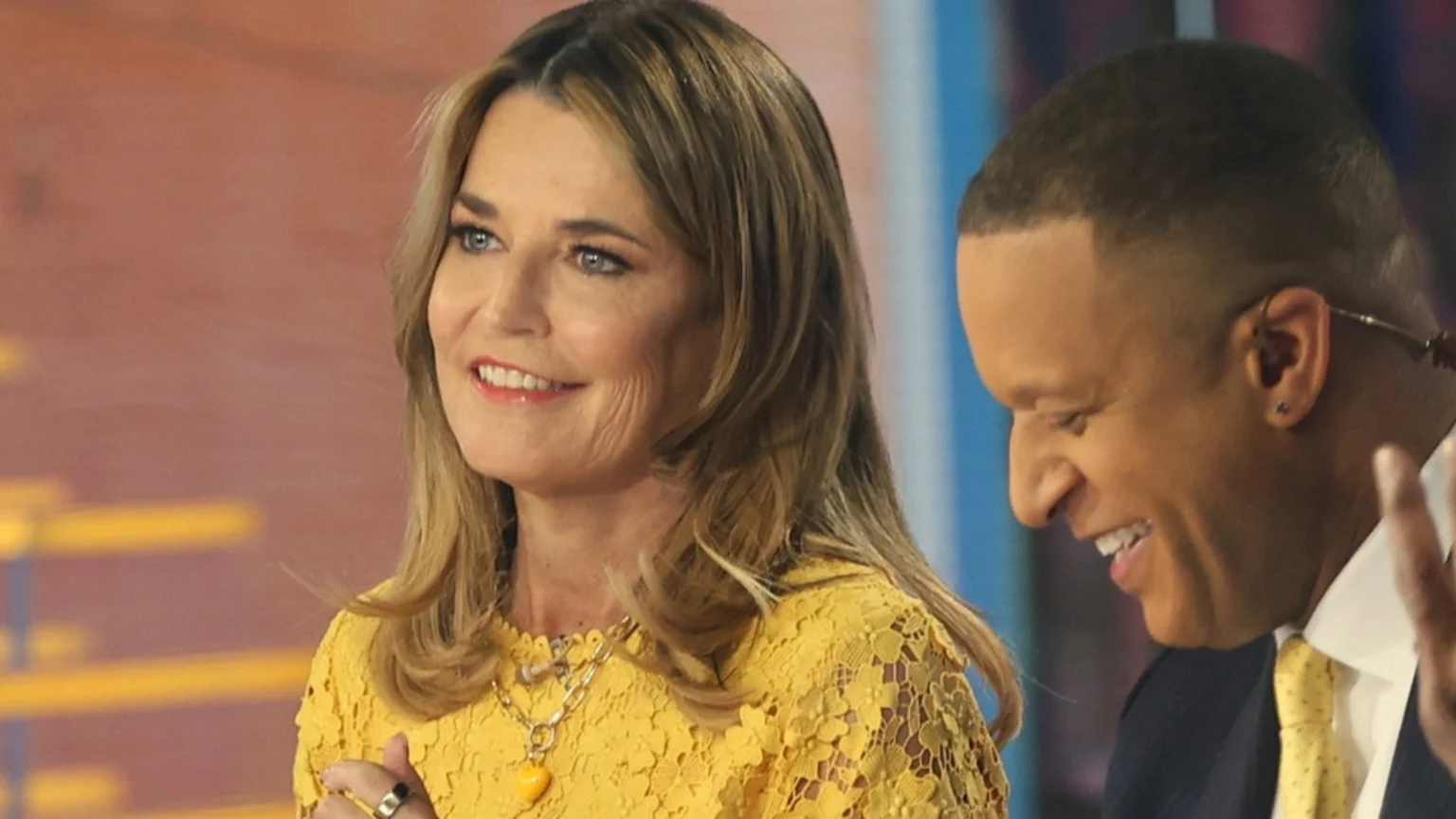 Savannah Guthrie, a woman with blonde hair, blue eyes, wearing a yellow lace top and a yellow heart necklace, smiles next to a man in a suit and yellow tie.