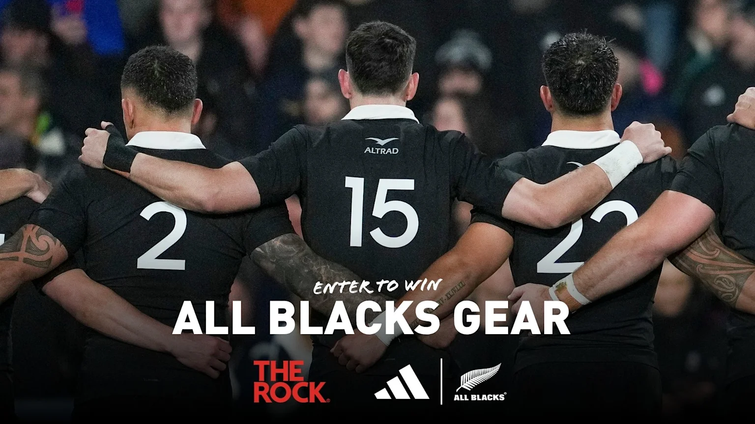 From behind, three All Blacks rugby players in black jerseys with numbers 2, 15, and 23 huddle together, with overlaid text "ENTER TO WIN ALL BLACKS GEAR" and sponsor logos.