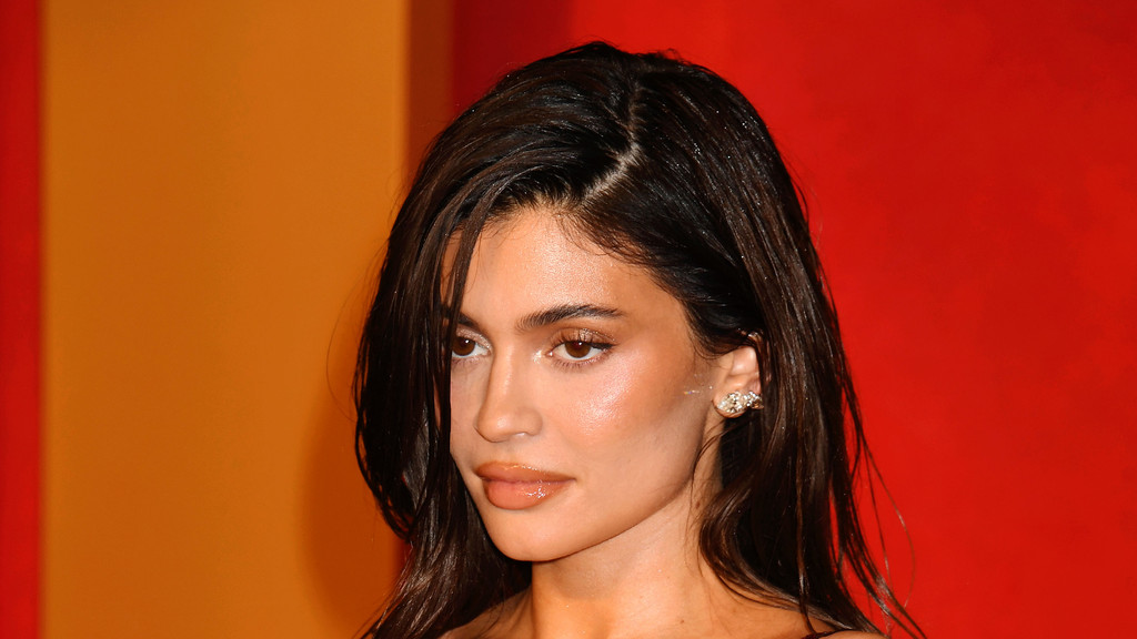 A woman with dark, glossy hair and subtle glitter, brown eyes, and full, glossy nude lips looks left in profile, wearing shimmering makeup and a decorative earring against an orange and red background.