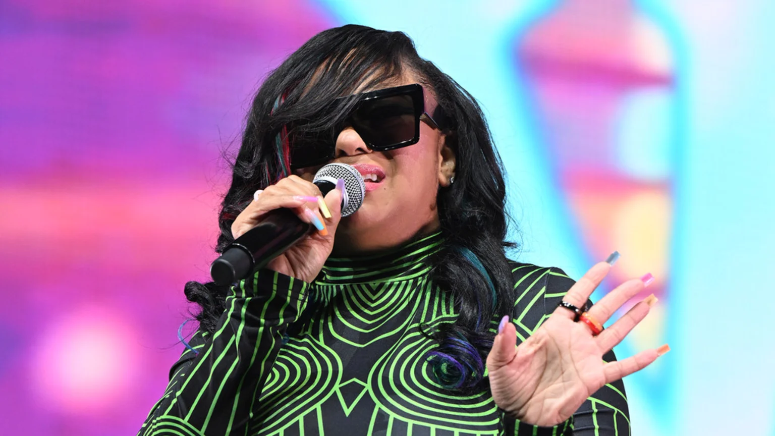 A woman with long dark hair highlighted with colorful streaks, wearing large sunglasses and a black-and-green patterned top, holds a microphone to her mouth with brightly painted nails, against a blurred colorful background.