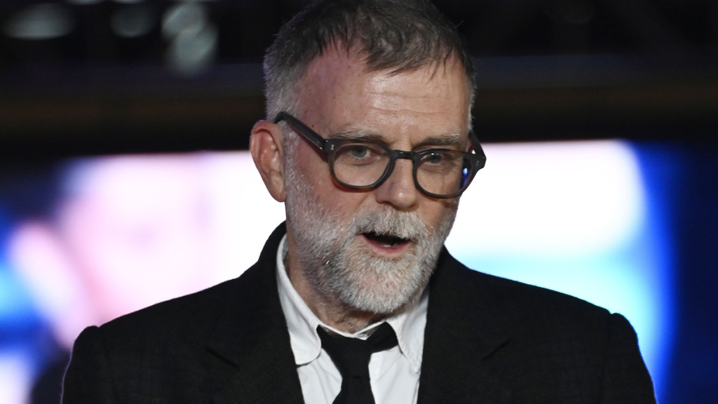 A man with gray hair and beard, wearing dark-framed glasses, a white shirt, black tie, and dark jacket, speaks against a blurry background.