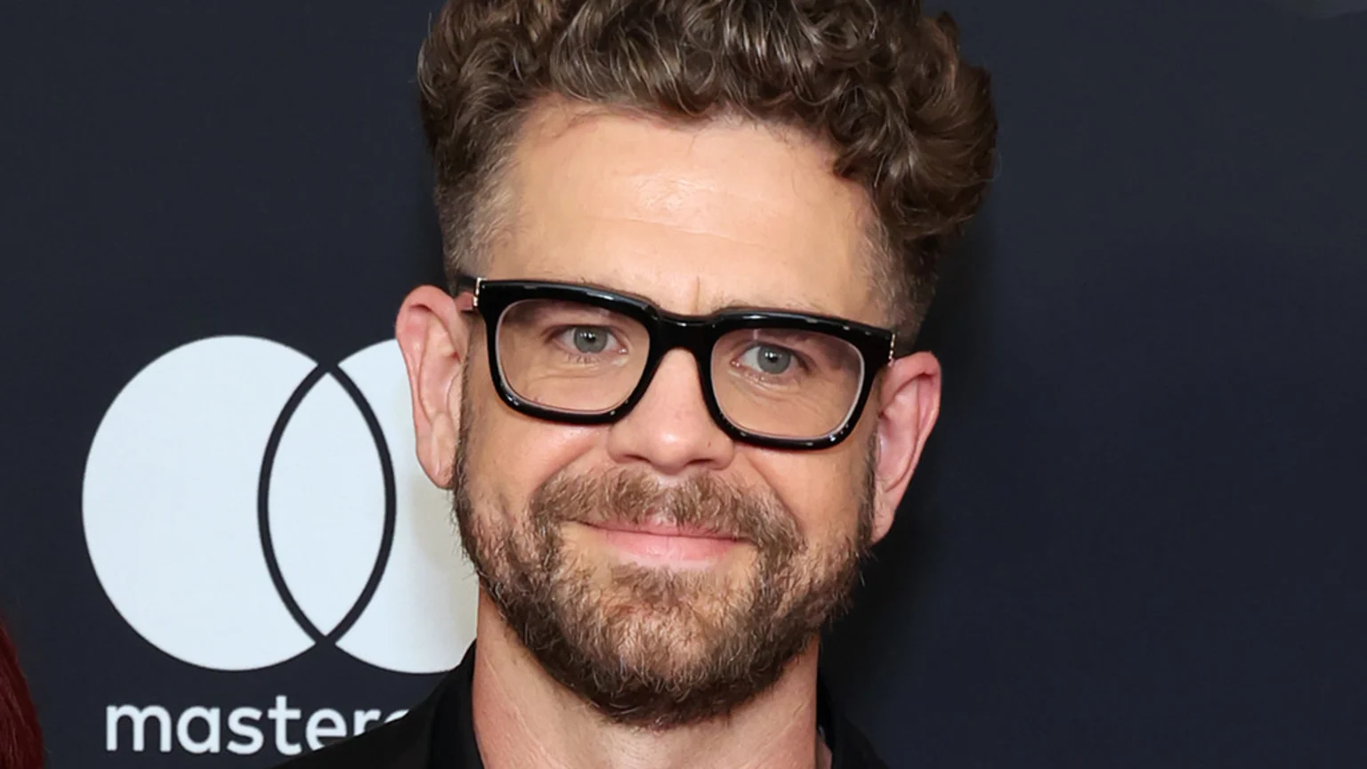A smiling Jack Osbourne, with curly brown hair, a beard, and black-framed glasses, is pictured against a dark background with a partial Mastercard logo.