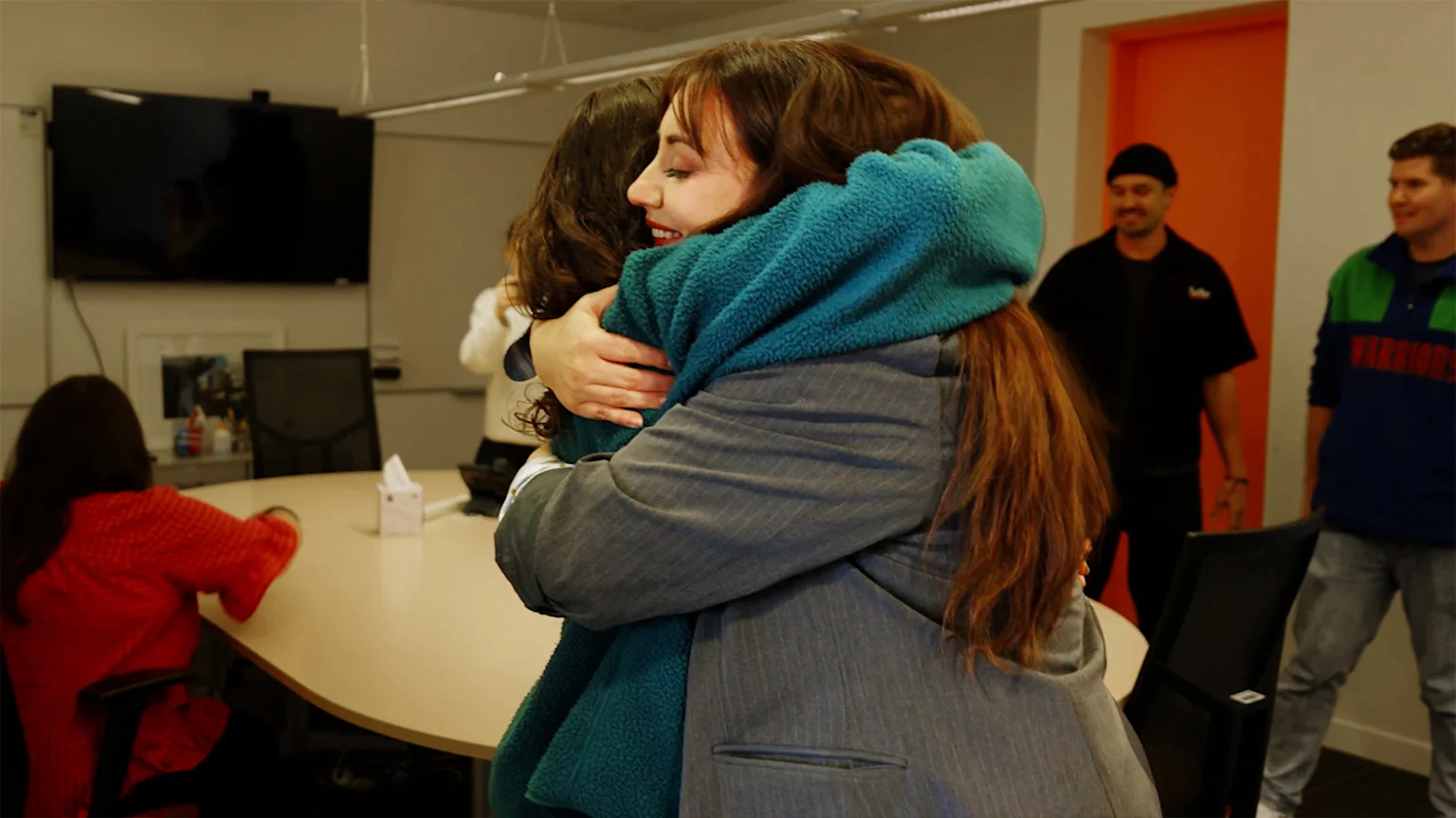 A woman with long brown hair, wearing a grey jacket and a teal fuzzy top, smiles while embracing another person, in what appears to be an office setting with other individuals visible.