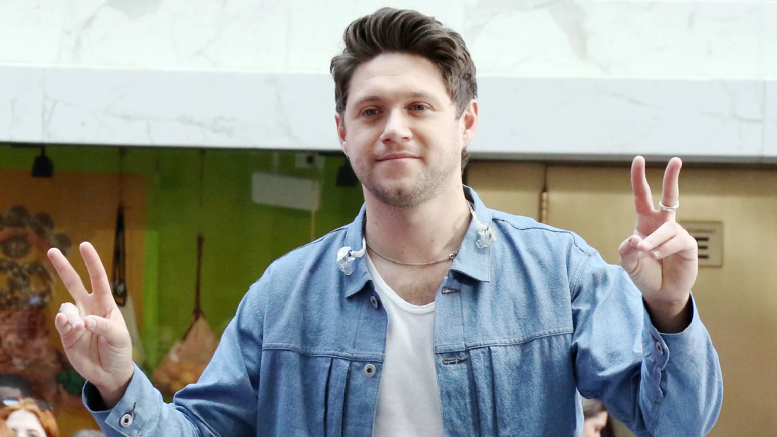 Niall Horan, wearing a denim jacket over a white t-shirt, smiles at the camera while holding up a peace sign with both hands.