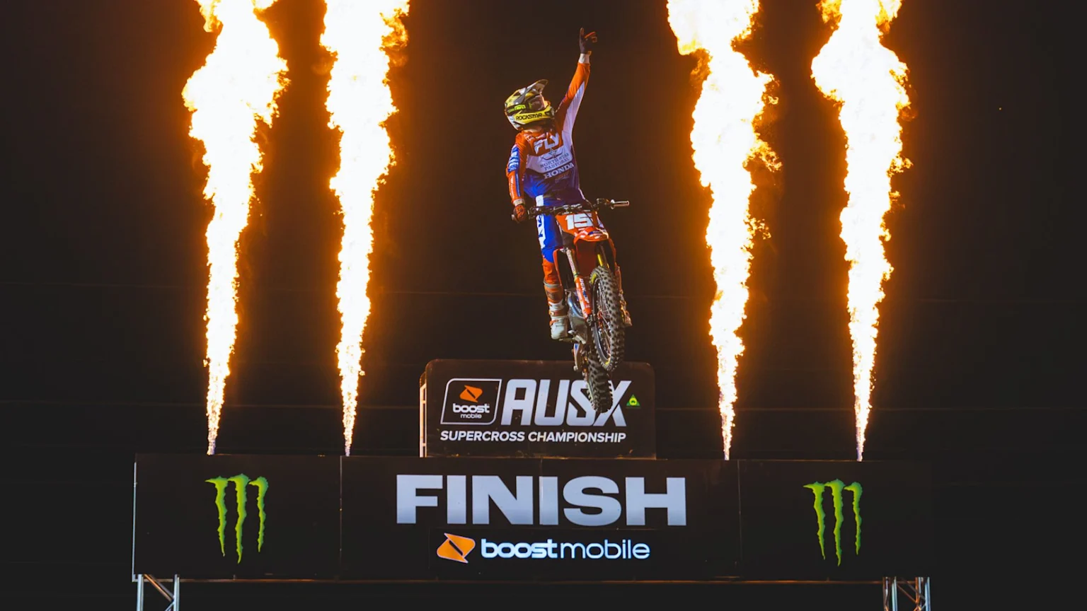 A motocross rider, number 15, with raised arm, jumps over the Boost Mobile AUSX Supercross Championship finish line, flanked by tall flames.