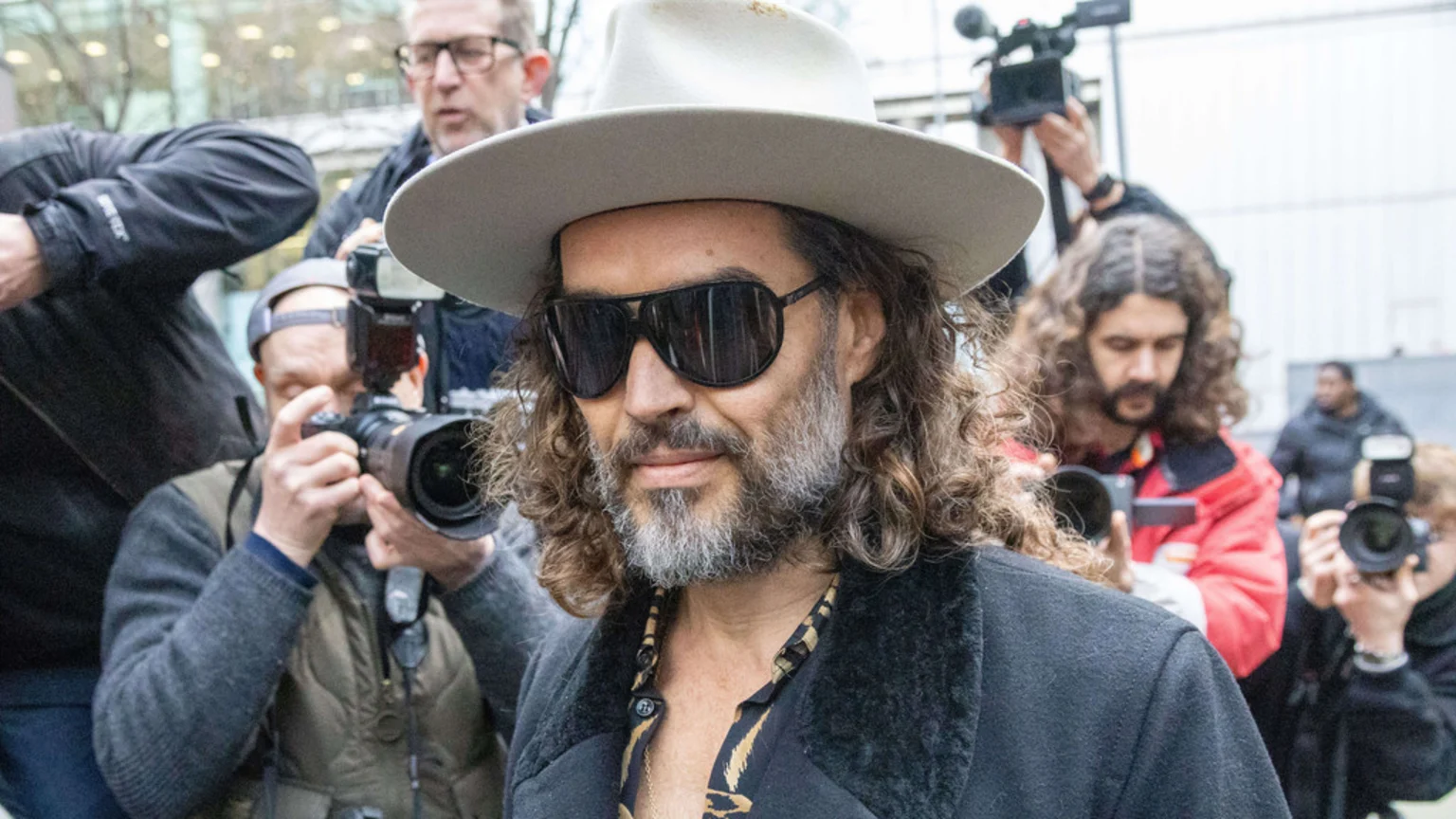 A man with a beard, long curly hair, sunglasses, and a wide-brimmed hat is surrounded by photographers and cameramen outdoors.