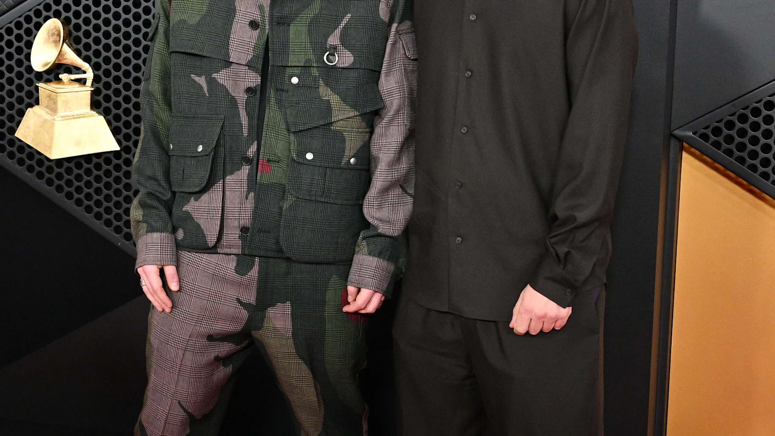 Ed Sheeran and Aaron Dessner stand on a red carpet. Sheeran wears a green and brown plaid suit, and Dessner wears a dark shirt, pants, beanie, and glasses, with Grammy awards in the background.