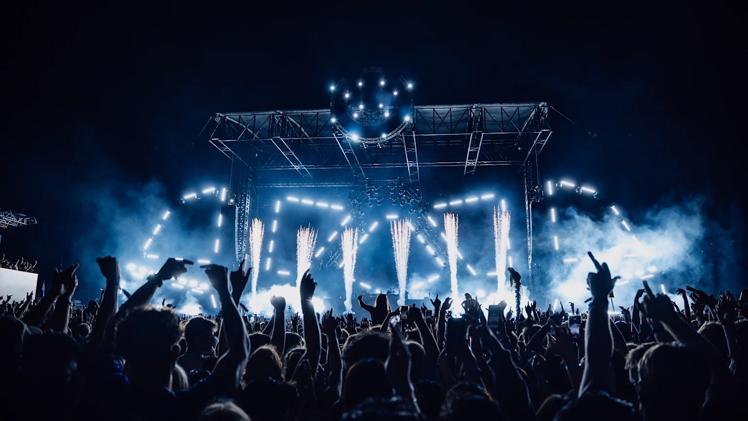 A massive crowd of silhouetted people with raised hands at a night concert, facing a stage lit with bright blue lights and pyrotechnics, under a large glowing disco ball.