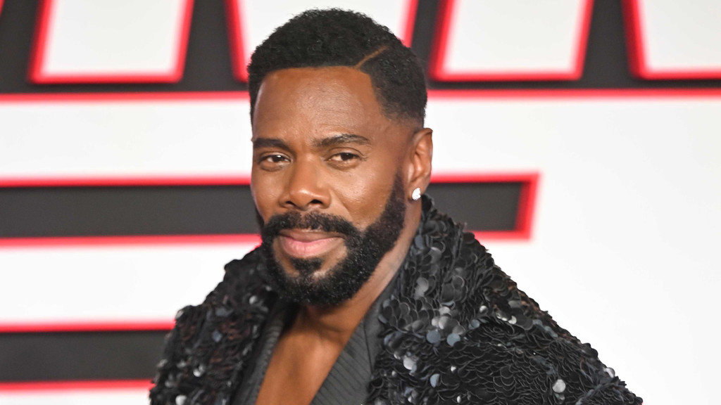A dark-skinned man with a beard and a short haircut smiles slightly, wearing a black sequined jacket and a diamond earring against a blurred white and red background.