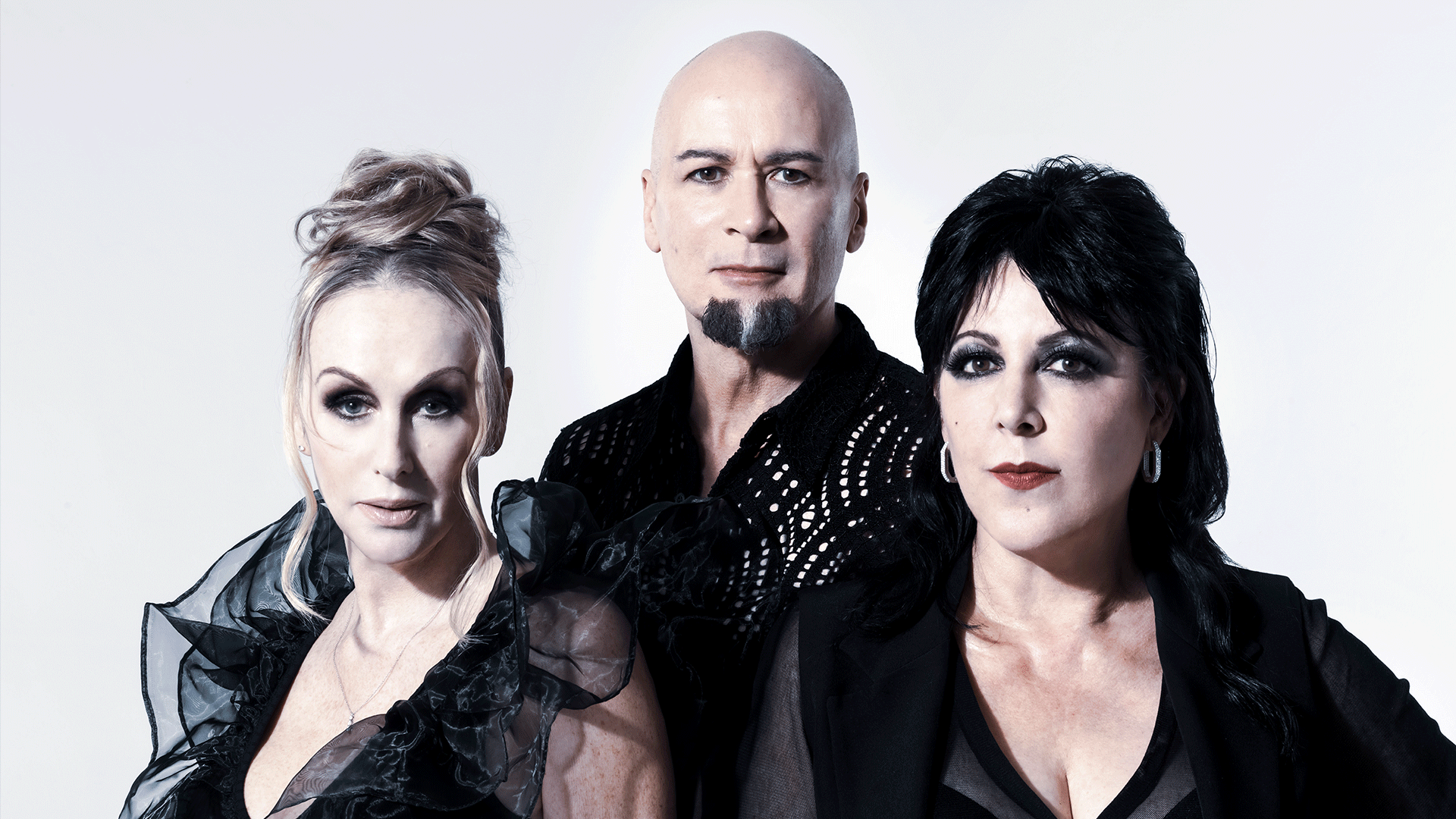 A group portrait shows two women and one man posing against a white background. The man in the center is bald with a goatee, flanked by a blonde woman on the left and a dark-haired woman on the right, all in black clothing.