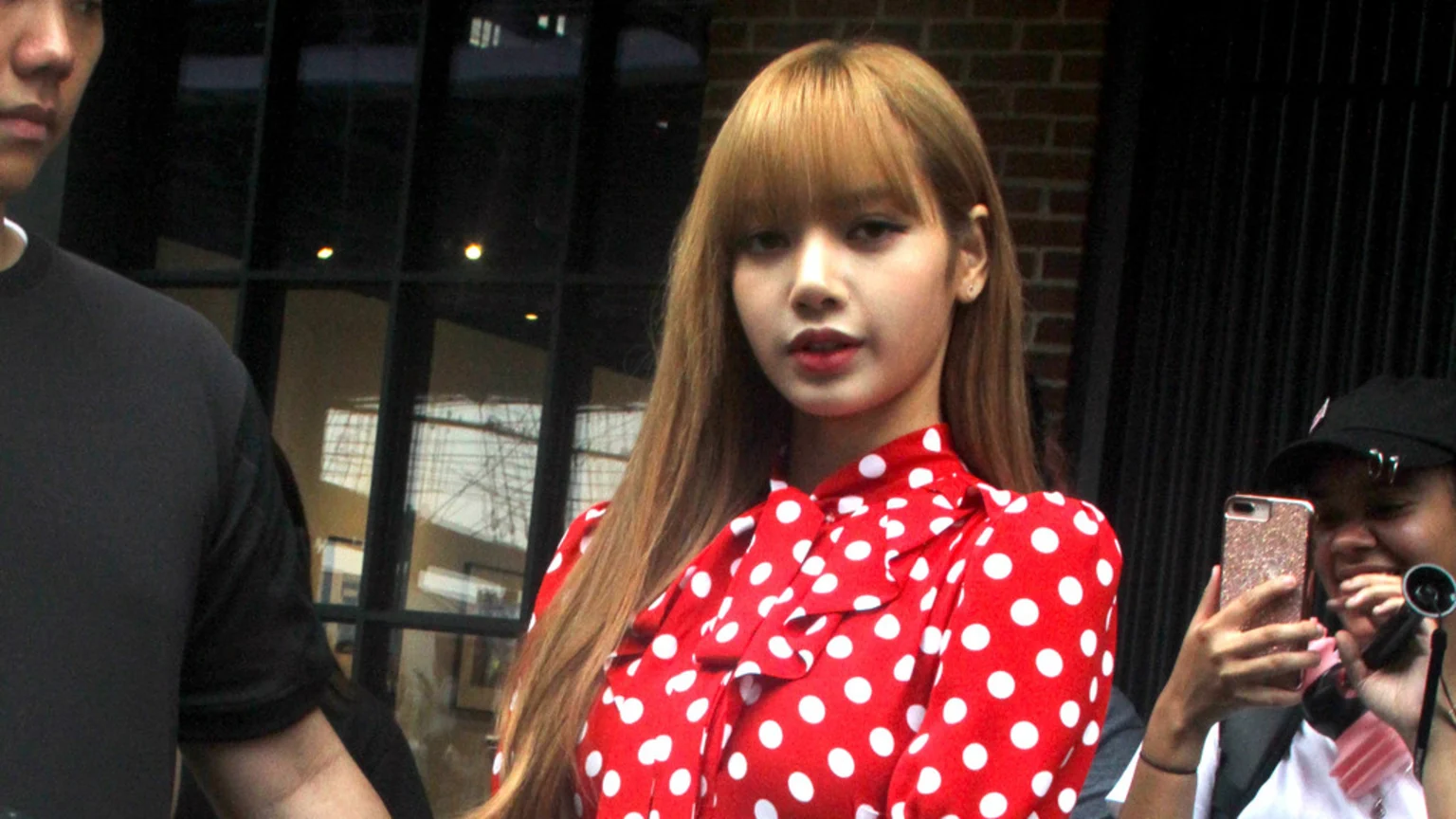 Blackpink's Lisa, with long brown hair and bangs, wears a red polka dot blouse and looks forward, while a person on the right holds a phone and laughs.