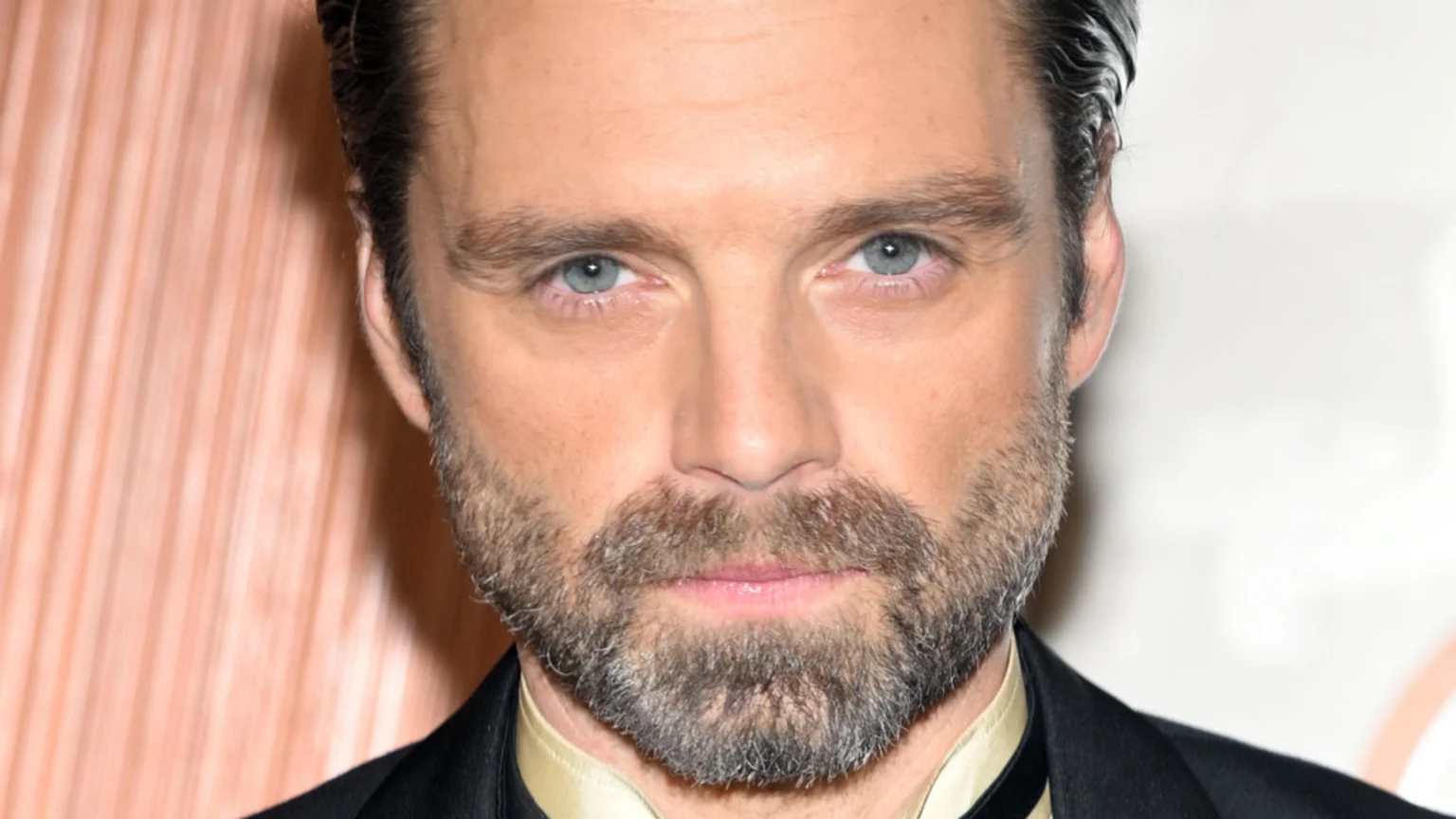 Close-up of actor Sebastian Stan looking at the camera, with light blue eyes, a beard, and dark hair, wearing a suit jacket and a light-colored shirt.