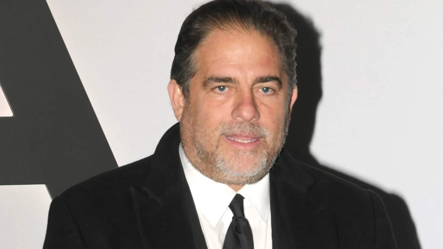 A close-up portrait of Harvey Weinstein, a middle-aged man with dark hair and graying beard, wearing a black suit and tie, posing with a neutral expression against a white and dark gray event backdrop with a partial letter 'A'.