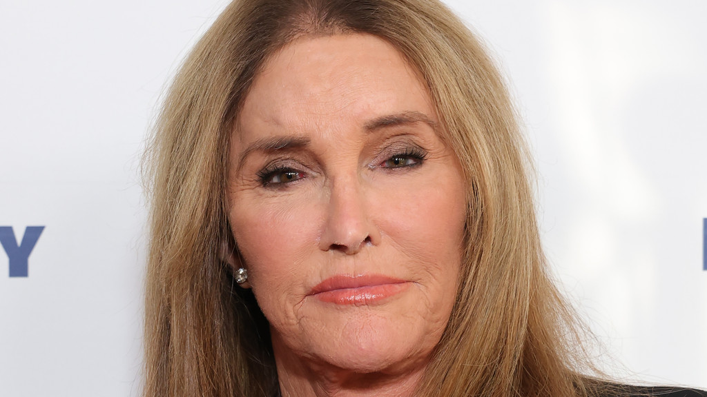 A close-up headshot of Caitlyn Jenner shows her with long blonde hair, makeup, and an earring, against a white background with blurred blue lettering.