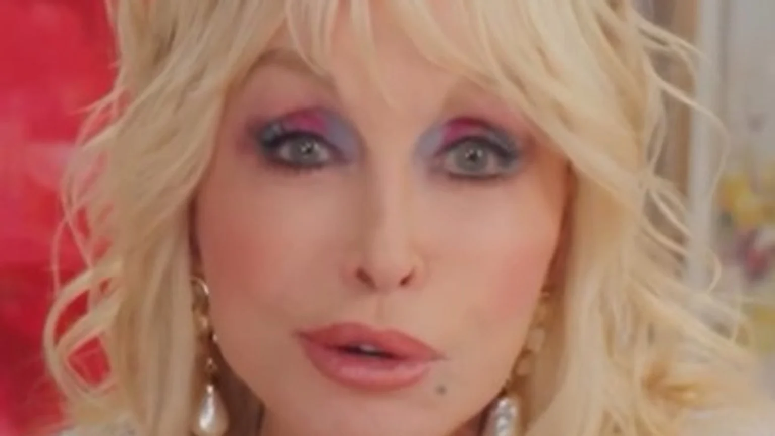 Close-up of Dolly Parton with blonde, wavy hair and distinctive blue and pink eyeshadow, looking directly at the viewer with slightly parted lips.