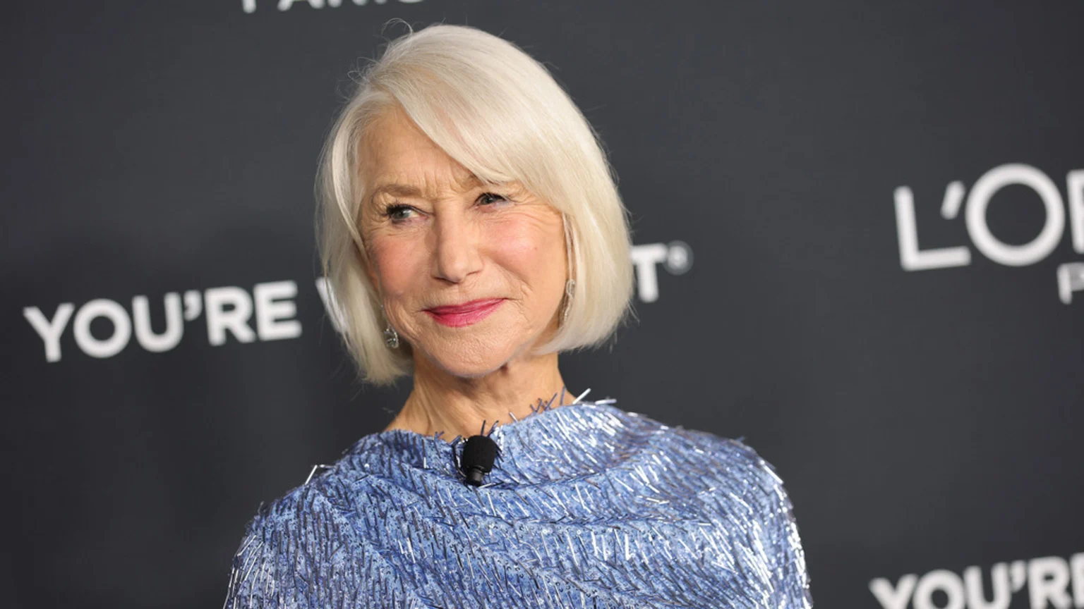 Close-up of Helen Mirren, smiling with short white hair, pink lipstick, and a blue sequined top with a microphone clipped to it, against a blurred dark background with white text.