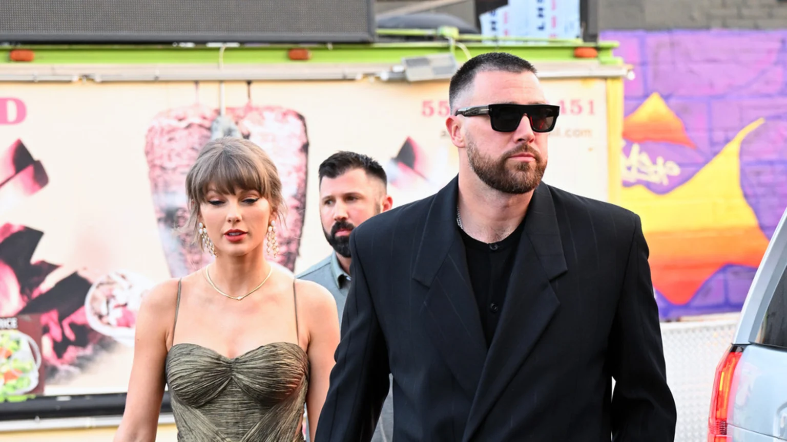 Taylor Swift, wearing a strapless, pleated gold dress and earrings, walks alongside Travis Kelce, who wears sunglasses and a black blazer, with a bearded man behind them.