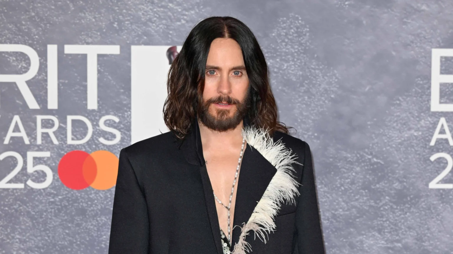 Jared Leto, with long dark hair and a beard, poses in front of a grey backdrop, wearing a black blazer with white feathers on the lapel and a long beaded necklace, with "BRIT AWARDS 25" and Mastercard logo visible.