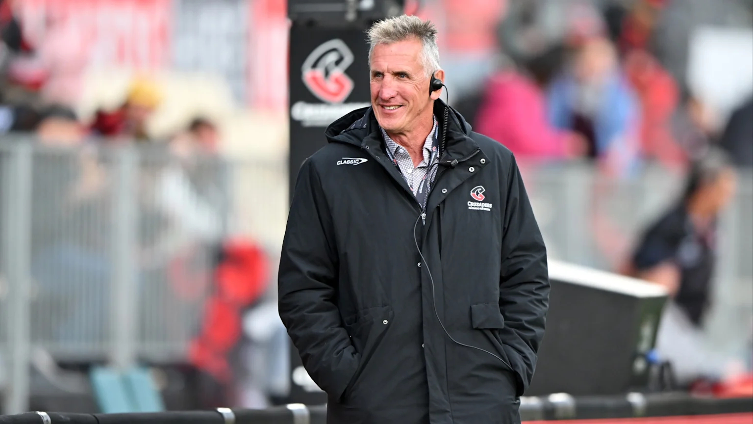 A smiling man with gray hair and an earpiece wears a black jacket with "CLASSIC" and "CRUSADERS" logos in a blurred stadium setting.