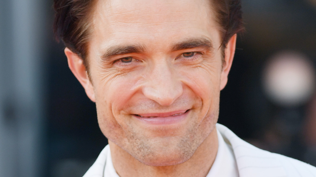 A close-up of actor Robert Pattinson smiling, with brown hair and light stubble, wearing a white collared shirt against a blurred dark background.