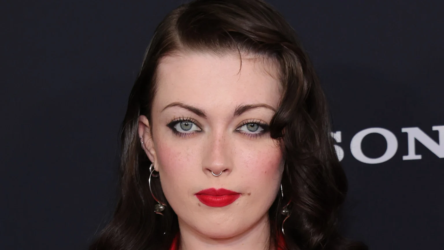 Close-up of a young woman with dark, wavy hair, prominent light eyes with dark eyeliner, red lipstick, and a silver septum piercing looking directly at the camera, against a dark background with the 'SONY' logo.