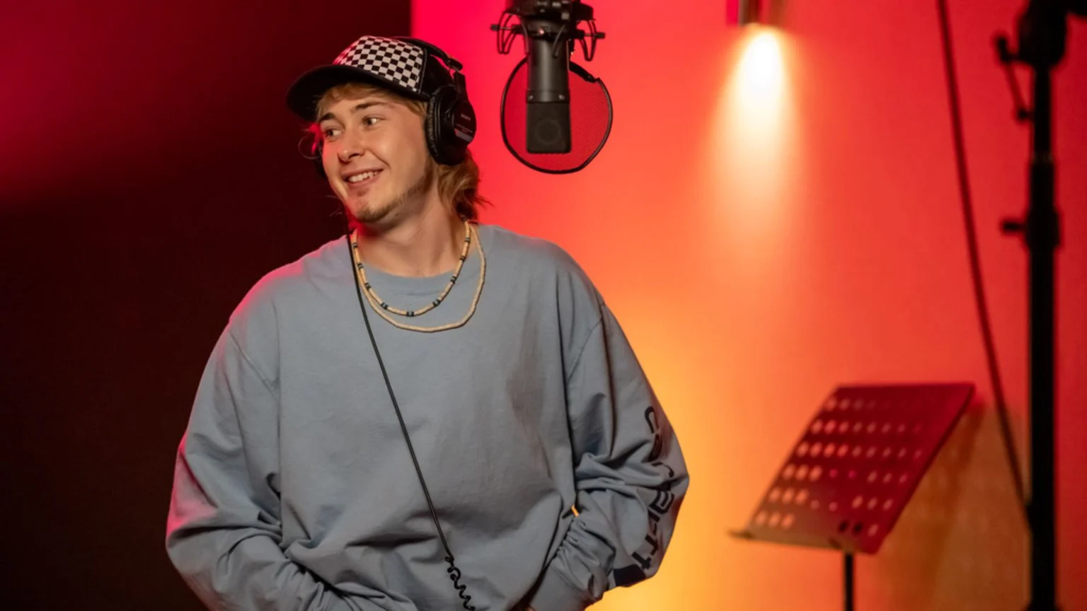 A smiling young man in a checkered cap and headphones stands in a red-lit recording studio, looking to the right, with a microphone in the background.