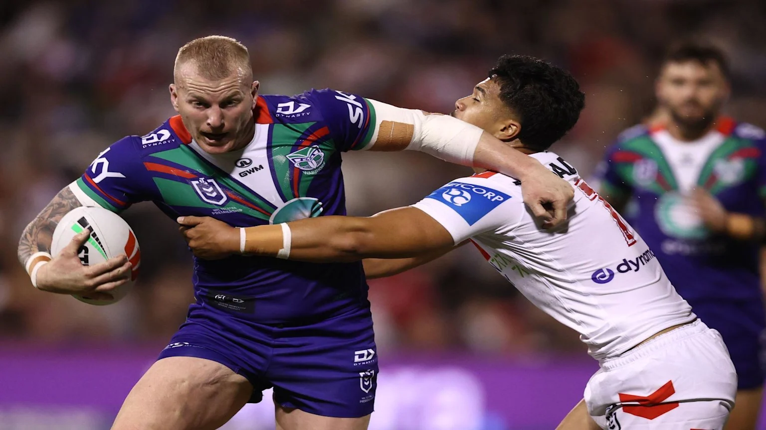 A blonde rugby player in a blue and green Warriors jersey runs with the ball while being tackled by an opponent in a white jersey.