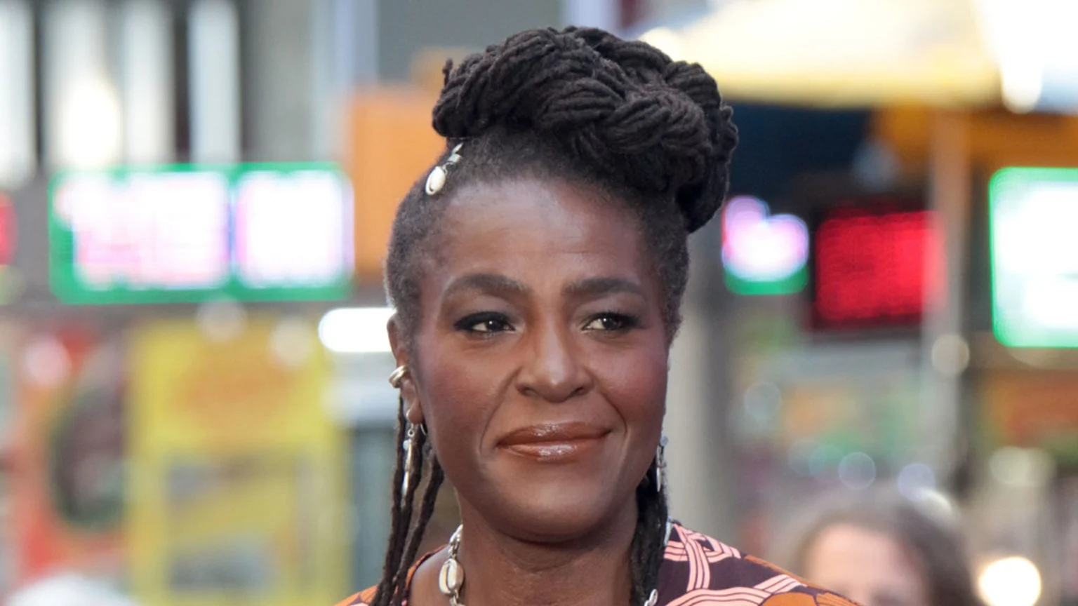 A close-up of a smiling woman with dark skin and dark dreadlocks styled in an elaborate bun, adorned with shell beads and earrings, with a blurred colorful background.