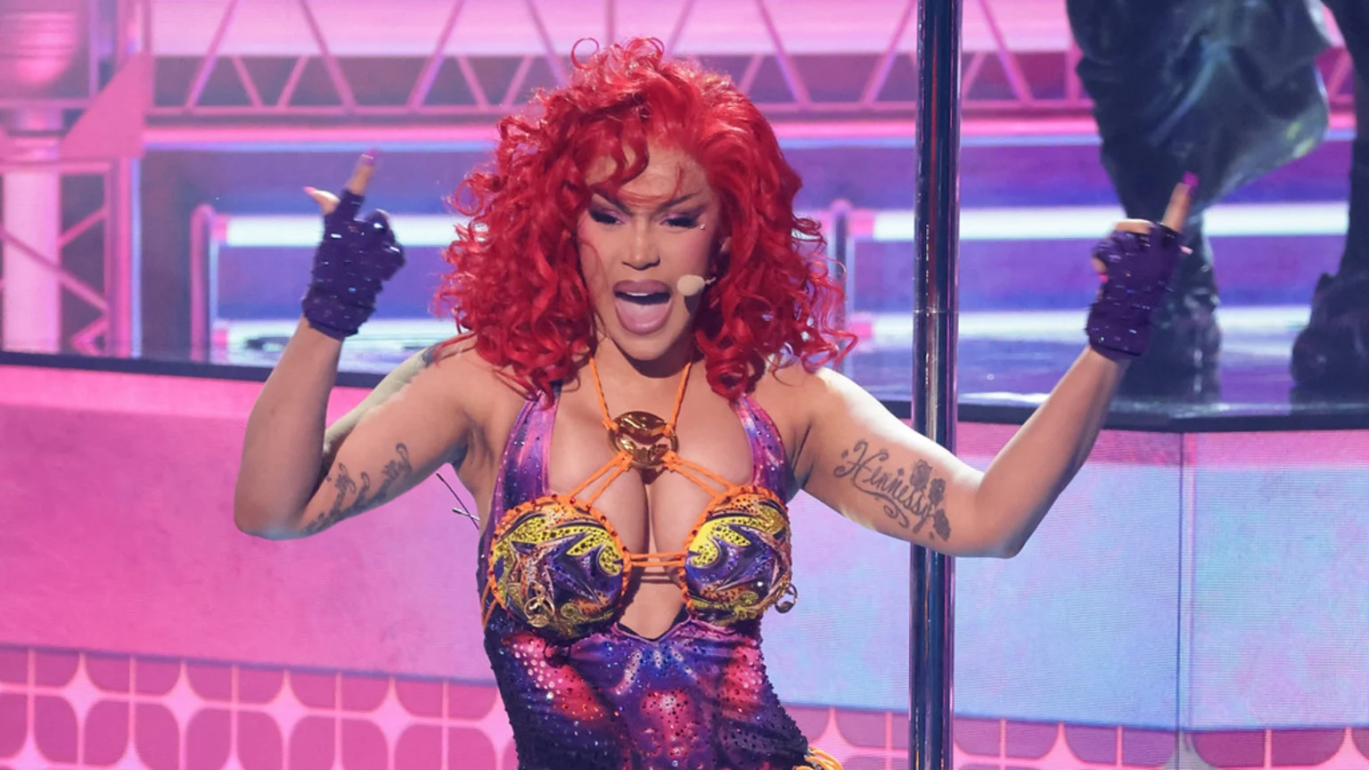 Cardi B on stage with bright red curly hair, wearing a sparkly, colorful outfit and purple gloves, pointing her fingers up while singing into a headset mic.