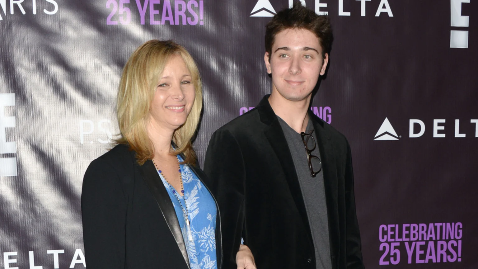 A blonde woman smiling in a black blazer stands arm-in-arm with a young man in a dark jacket and glasses on his collar, posing in front of a dark backdrop with "DELTA" and "CELEBRATING 25 YEARS!" visible.