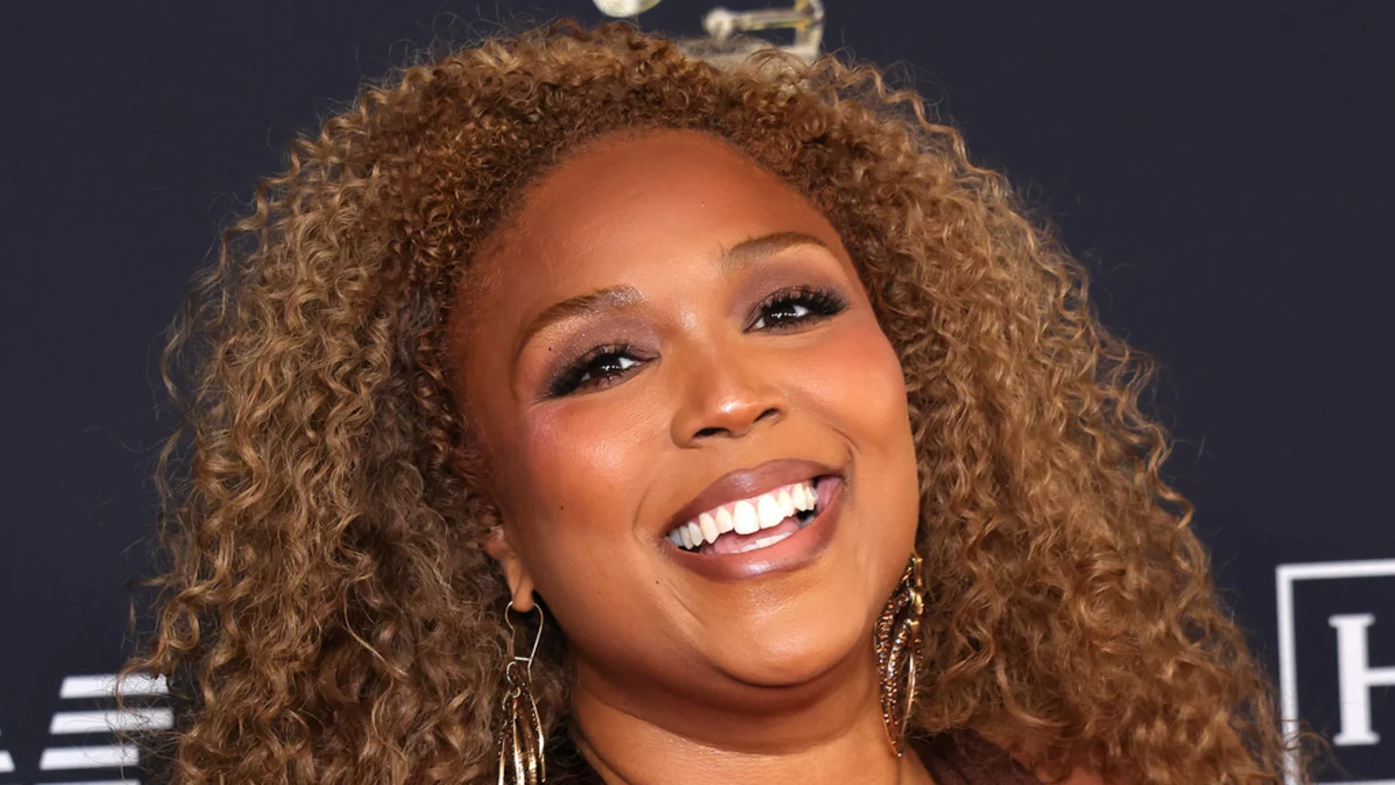 A woman with curly light brown hair smiles broadly, showing white teeth, wearing gold earrings and warm-toned makeup against a dark background.
