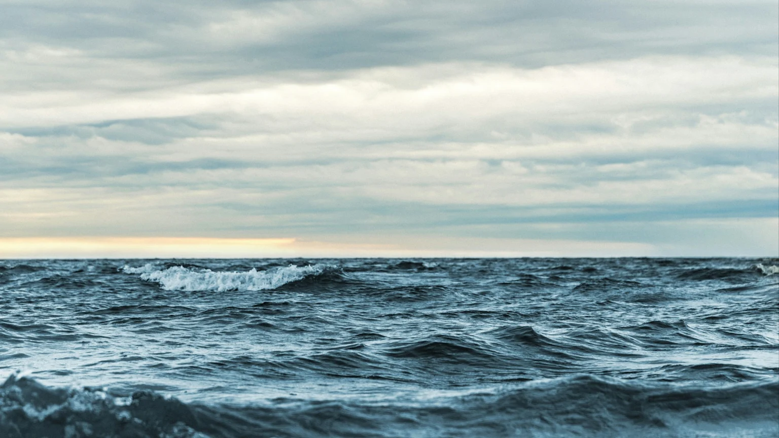 Dark blue ocean water with numerous waves and some whitecaps extends to a cloudy horizon with a faint golden glow, beneath a light grey and white sky.