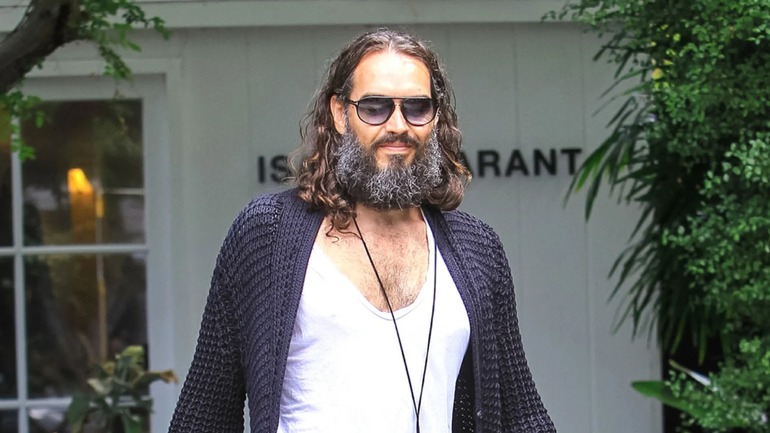 Russell Brand, with long brown hair, a grey beard, and sunglasses, smiles faintly while wearing a white t-shirt and dark open cardigan, standing outdoors.