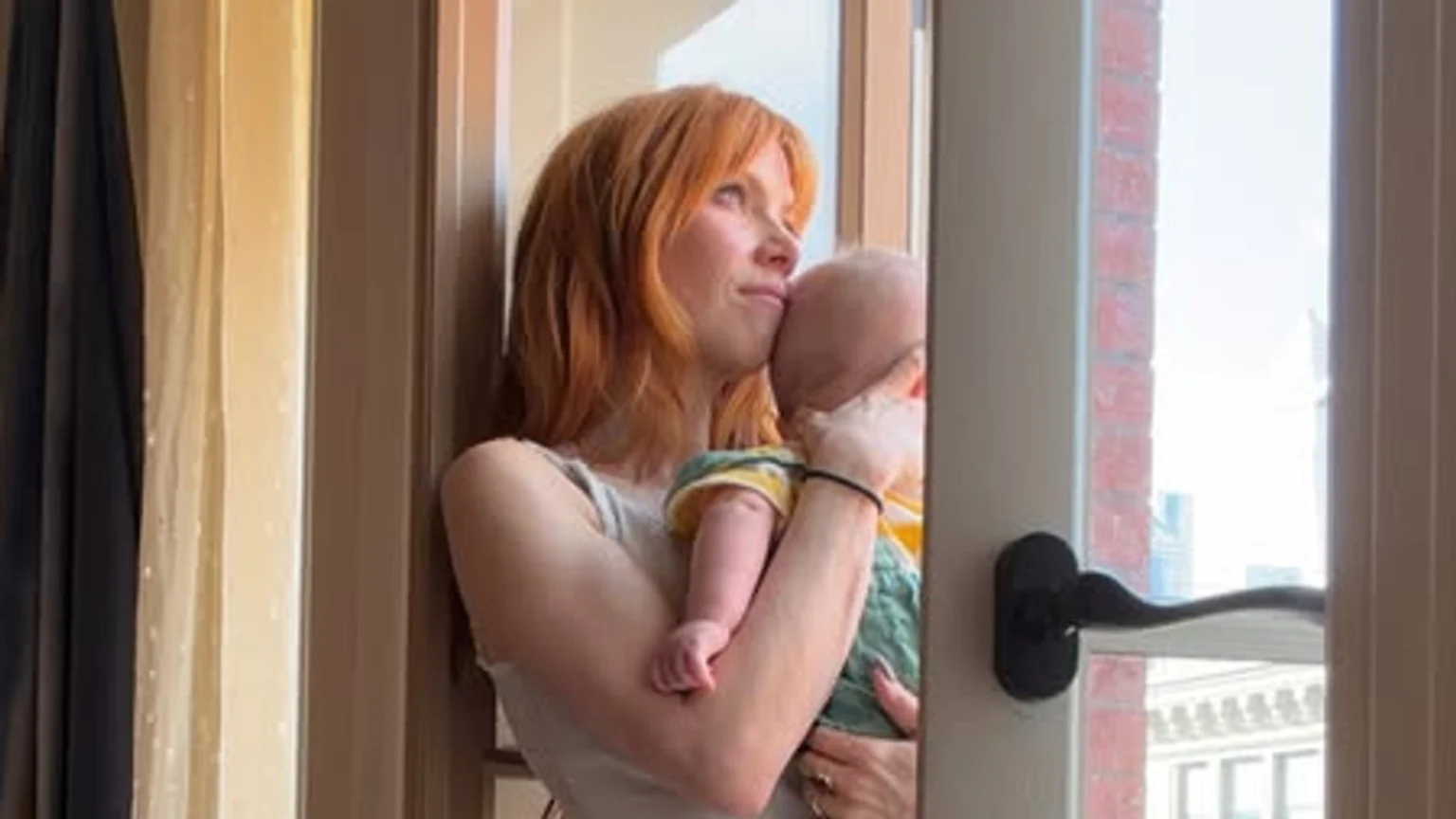 A woman with orange hair holds a baby, looking up and away with her cheek resting on the baby's head, in a sunlit indoor setting with a partial view of a city outside.