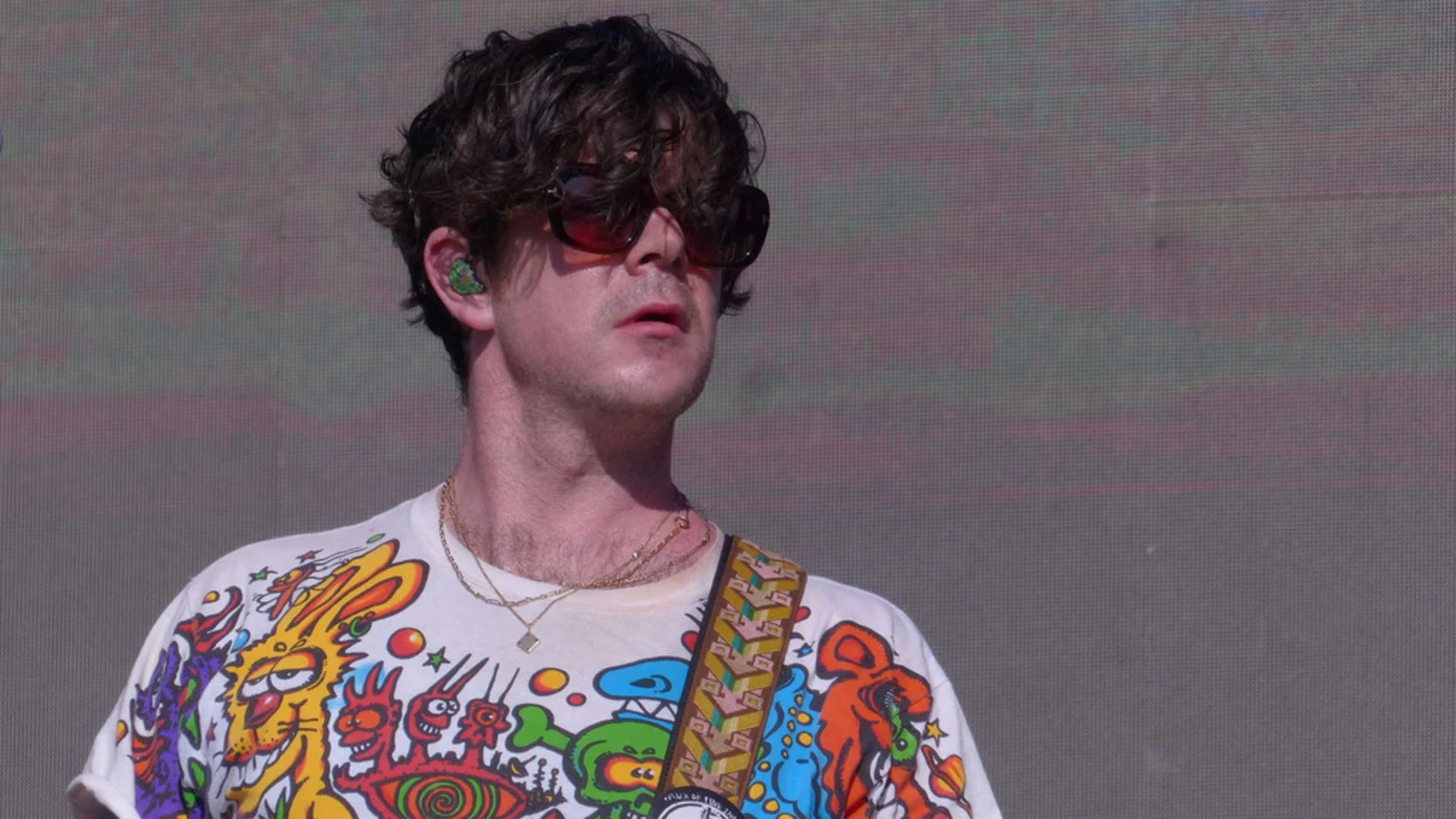 A man with dark curly hair and sunglasses, wearing a white t-shirt with colorful cartoon graphics, gold necklaces, and a guitar strap, looks up with a pixelated screen behind him.