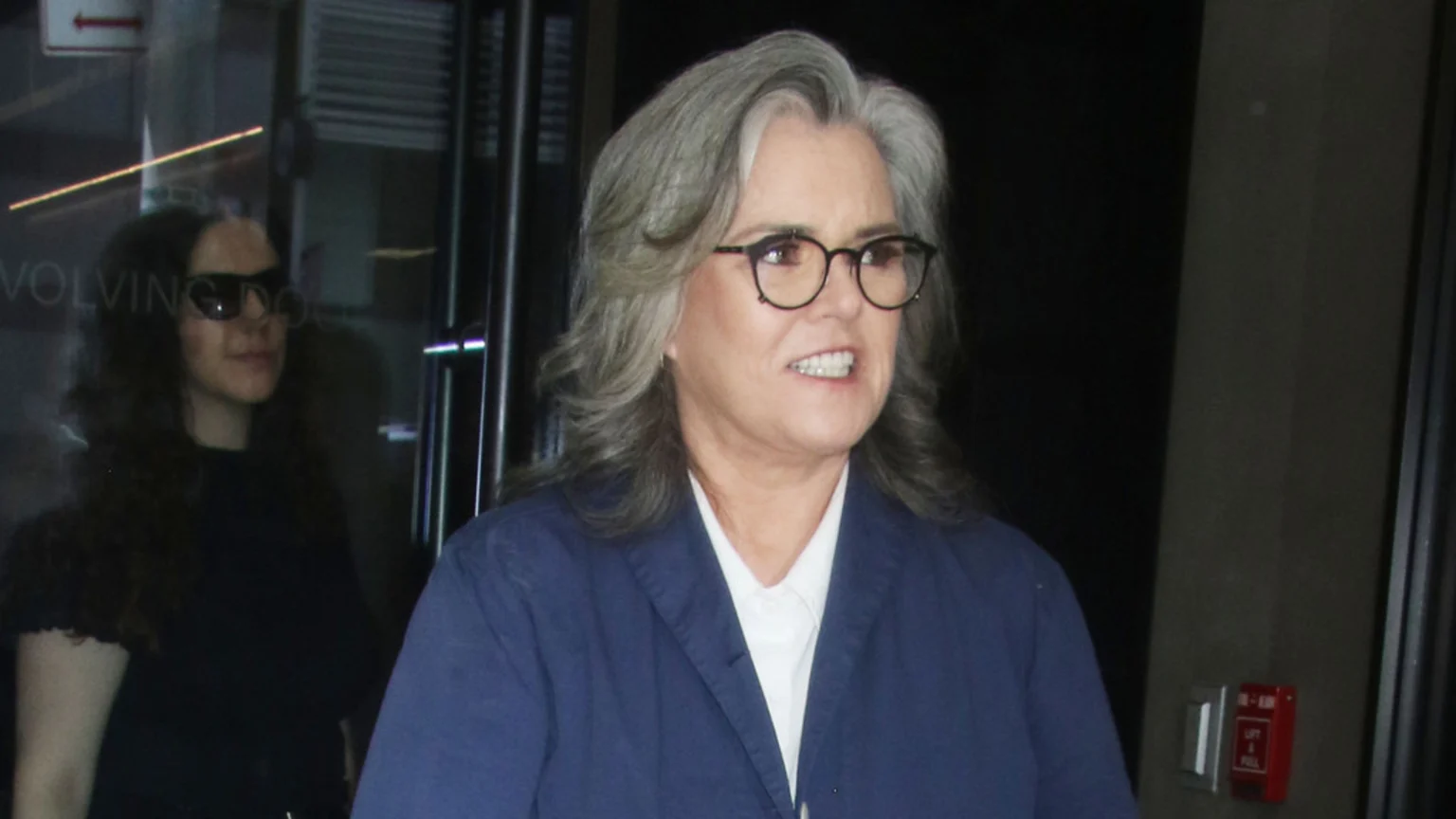 Rosie O'Donnell, with grey hair and glasses, wears a blue jacket over a white shirt, mouth slightly open, with a woman in sunglasses partially visible in the blurry background.
