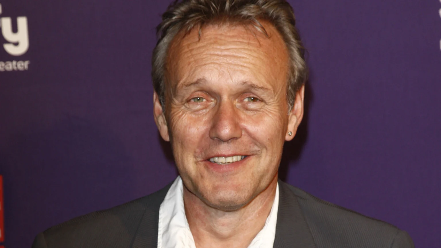 A smiling middle-aged man with greyish-brown hair, green-brown eyes, a white shirt, dark jacket, and a silver stud earring, against a blurry purple background with "Syfy" text.