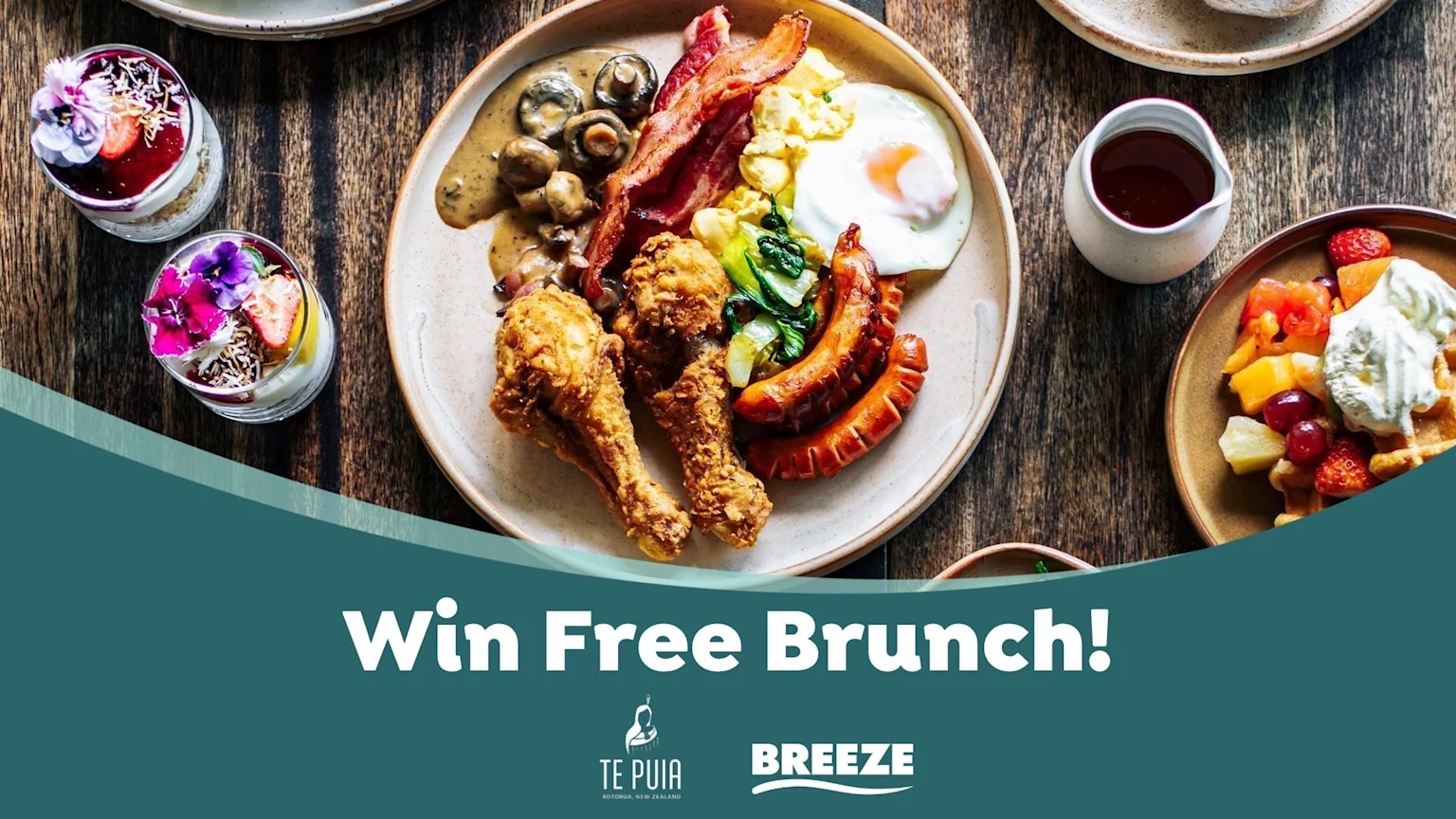 A wooden table with a large brunch platter of fried chicken, bacon, eggs, sausages, and mushrooms, plus fruit parfaits and waffles, below a banner reading 'Win Free Brunch!'.
