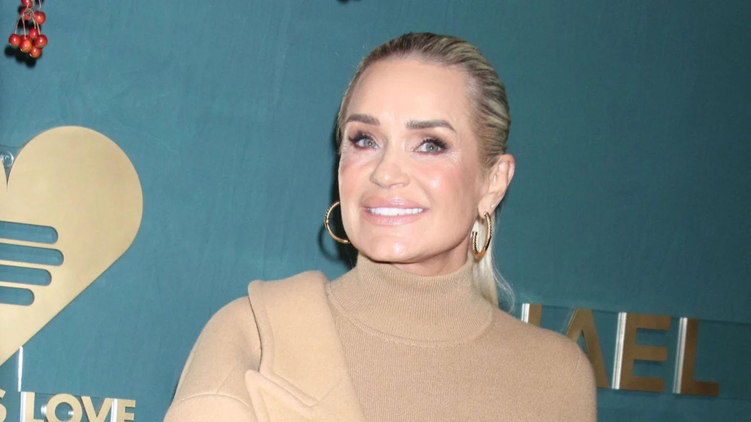 Yolanda Hadid, a blonde woman with her hair pulled back, smiles while wearing a camel turtleneck and gold hoop earrings against a teal background with a gold heart logo.