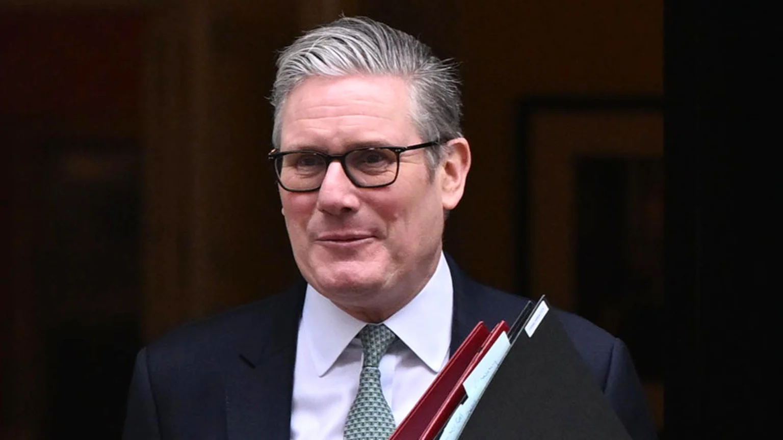 Keir Starmer, a man with gray hair and glasses, wearing a suit and patterned tie, smiles slightly while holding several folders.