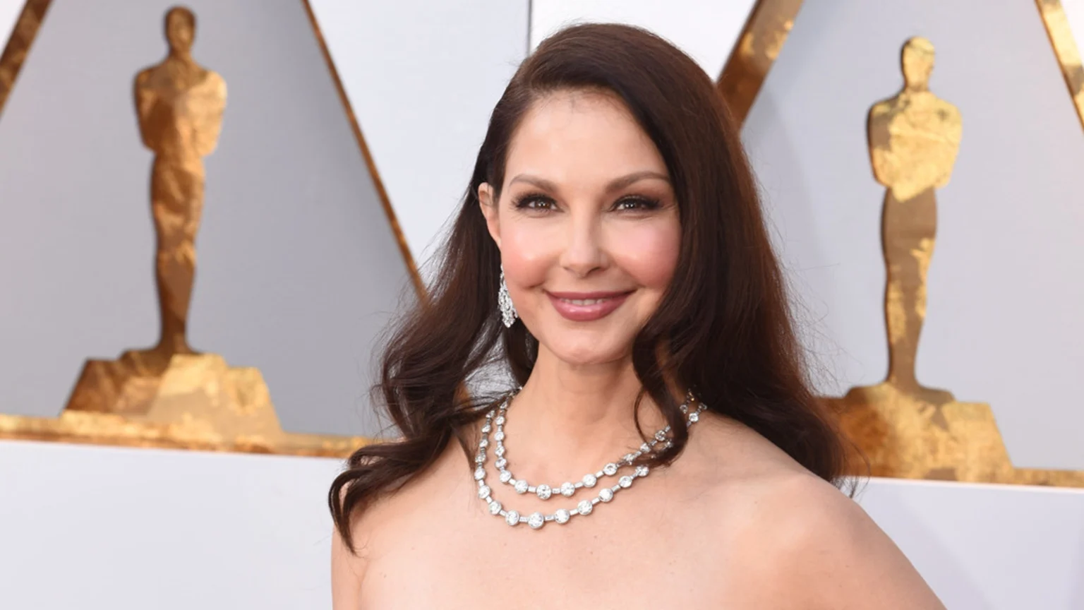 Smiling Ashley Judd, with long dark brown hair and wearing a strapless gown, accessorized with sparkling diamond earrings and a two-strand necklace, stands in front of blurry gold Oscar statuettes.
